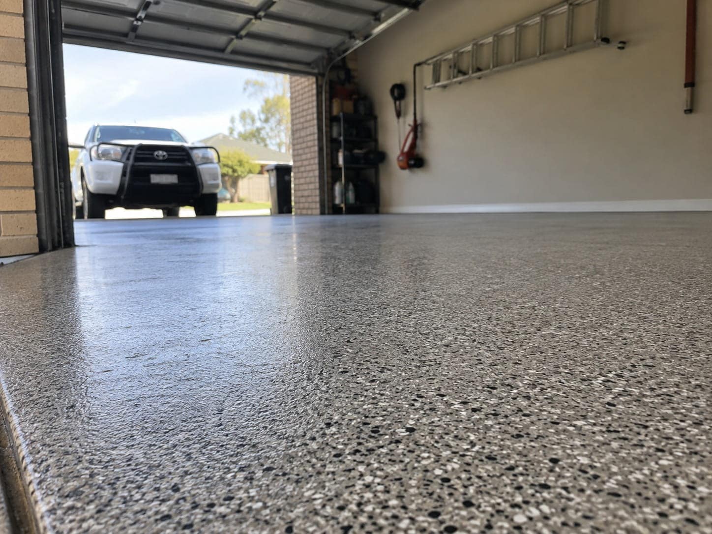 Polished speckled garage floor at a Mandurah home, photographed low with the roller door open and a ute parked on the driveway beyond
