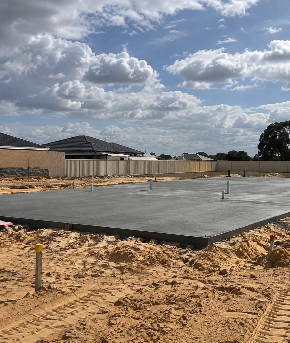 Freshly poured shed slab with service risers standing on sandy Mandurah ground, formed edge and neighbouring rooflines behind