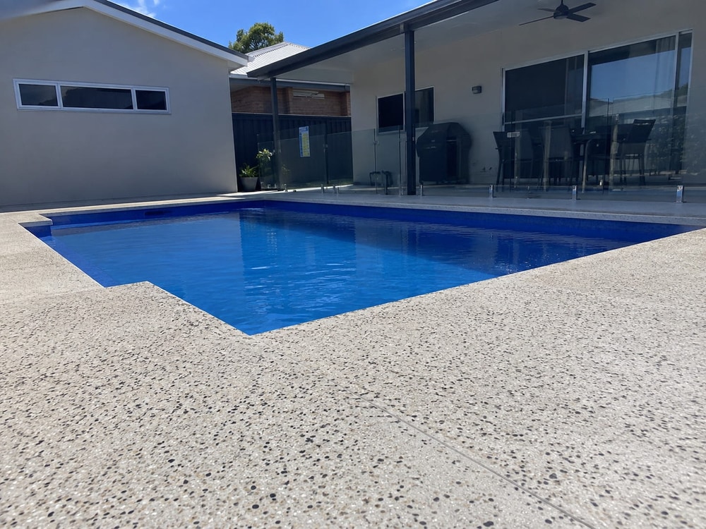 Pale exposed aggregate pool surround beside a blue pool with glass fencing